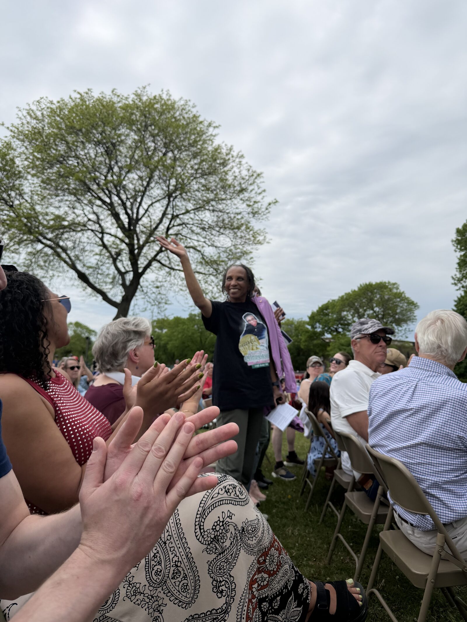 A family member of Anna Robertson stands and waves with a wide smile, wearing a t-shirt commemorating Robertson, as surrounding audience members applaud during the Memorial Day tribute